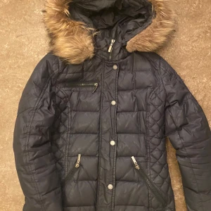 Svart dunjacka från Rockandblue - Säljer en svart dunjacka från Rockandblue med snygg pälsdetalj på huvan. Jackan har flera praktiska dragkedjor och en cool patch på ärmen. Perfekt för kalla vinterdagar!  den har ett litet hål på armen men den syns knappt. 