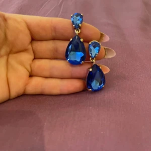 Blå glittriga örhängen - Snygga örhängen med stora, droppformade blå stenar som glittrar i ljuset. Perfekta för en fest eller en speciell kväll. De har en elegant design med en mindre sten ovanför den större, vilket ger en lyxig känsla. Passar perfekt till en klänning eller en stilren outfit.