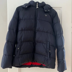 Mörk blå dunjacka från Gant - Säljer en mörkblå dunjacka från Gant i mycket bra skick. Jackan har en dragkedja framtill och en mysig huva. Perfekt för kalla vinterdagar. Den är vadderad och håller dig varm och skön. Passar både till vardags och lite finare tillfällen.