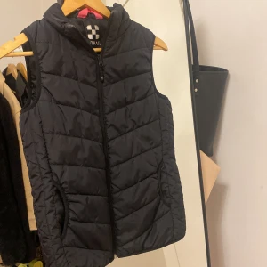 Svart dunväst från Trail - Säljer en svart dunväst från Trail i mycket bra skick. Västen har en dragkedja framtill och är perfekt för höst och vinter. Passar storlek 36