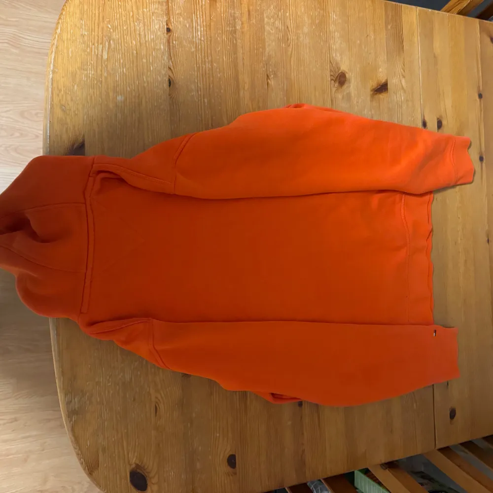 Säljer en snygg orange hoodie från Tommy Hilfiger med vit text och logga på framsidan. Den har en klassisk känguruficka och justerbar huva med vita snören. Perfekt för en avslappnad stil och passar bra till höst och vår.. Tröjor & Koftor.