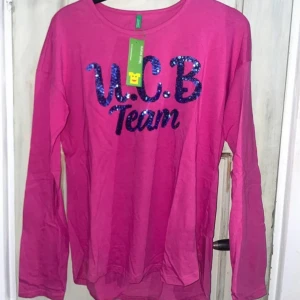 Ny Benetton rosa långärmad tröja med paljetter - Säljer en superfin rosa långärmad tröja från United Colors of Benetton. Den har en cool paljetttext 'U.C.B Team' på framsidan som verkligen poppar! Tröjan är i nytt skick med etikett kvar.  Barnstorlek 3xl, 13-14 år, 170.  Orginalpris ca 170kr. 💖