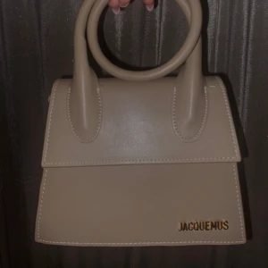 Beige handväska från Jacquemus - Säljer en superfin beige handväska från Jacquemus. Den har en stilren design med rundade handtag och är perfekt för både vardag och fest. Väskan är i mycket bra skick och har en elegant look som passar till alla outfits.