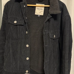 Svart manchesterjacka från Jack & Jones - Säljer en snygg svart manchesterjacka från Jack & Jones. Den har en mysig fleecefoder på insidan och knappar framtill. Perfekt för höst och vår, med en cool och avslappnad stil. Jackan har två bröstfickor med knappar och en klassisk krage.