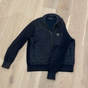Mörkblå jacka från J.Lindeberg - Säljer en snygg mörkblå jacka från J.Lindeberg i storlek S. Jackan har en dragkedja framtill och är quiltad på framsidan med ribbade ärmar och krage. 