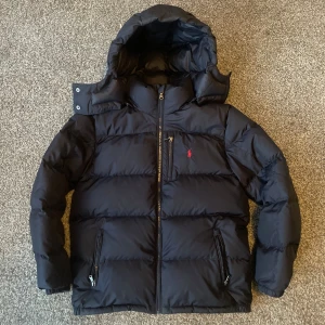  dunjacka från Ralph Lauren - Säljer en snygg  dunjacka från Ralph Lauren. Jackan har en huva och dragkedja framtill, samt två praktiska fickor med dragkedjor. Perfekt för kalla vinterdagar. Den är i mycket bra skick och passar både till vardags och lite finare tillfällen. Storleken är L ungdom passar xs