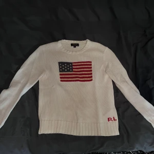 Vit stickad tröja från Ralph Lauren - Säljer en snygg vit stickad tröja från Ralph Lauren med en amerikansk flagga på bröstet. Tröjan har långa ärmar och är i mycket bra skick. Perfekt för höst och vinter, och den är supermysig att ha på sig. Passar både till vardags och lite finare tillfällen.