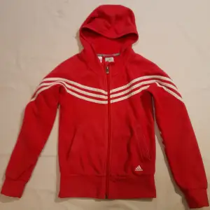 Säljer en snygg mörkrosa huvtröja från Adidas i storlek 140. Den har de klassiska tre vita ränderna längs ärmarna. Tröjan är i fleece, har en dragkedja framtill och en bekväm huva. Perfekt för både träning och vardag. Den är i mycket bra skick och redo för nya äventyr!