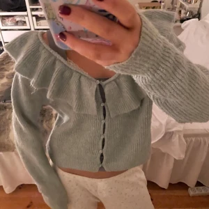 Ljusgrön stickad kofta med volang - Superfin ljusgrön stickad kofta med volangdetaljer längs halsringningen. Den har långa ärmar och knäpps med knappar. Perfekt mysig passar till allt