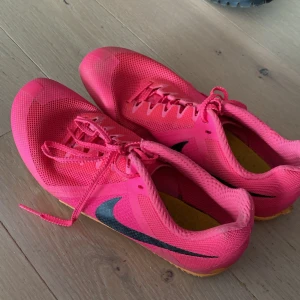 Rosa löparskor från Nike - Säljer ett par rosa löparskor från Nike i bra skick. De har en svart swoosh-logga och är utrustade med spikar under sulan för extra grepp. Perfekta för löpning på bana. Skorna har snörning och är gjorda i ett lätt syntetmaterial. Man får en grön ”skruvmejsel” för att ta bort och sätta på spikarna på skorna.