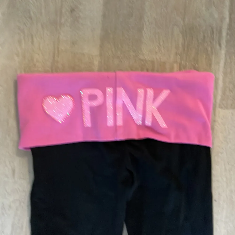 Säljer ett par svarta byxor med en bred rosa midja . De har en hjärtdetalj och texten 'PINK' i glittriga bokstäver. Byxorna är i bootcut-stil och ser superbekväma ut för en avslappnad dag hemma eller på stan.. Jeans & Byxor.