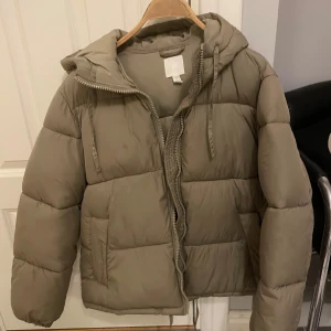 Beige pufferjacka med huva - Säljer en snygg beige pufferjacka i mycket bra skick. Jackan har en praktisk huva och dragkedja framtill. Perfekt för kyliga höst- och vinterdagar. Den är varm och bekväm, idealisk för både vardag och utomhusaktiviteter. 