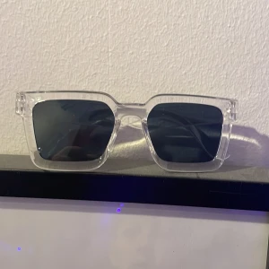 Transparenta solglasögon med svart lins - Säljer ett par stilrena solglasögon med transparent båge och svarta linser. Perfekta för en modern look och passar till alla outfits. De har en fyrkantig form som ger en cool och trendig stil. Perfekta för både vardag och fest!