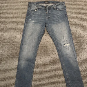 Blå jeans från Jack & Jones - Säljer ett par snygga blå jeans från Jack & Jones i modellen Glenn som är väldigt populära!  Storlek 34/32. Jeansens slitningar gör dom extremt snygga även fast att det är hål vid slitningen vid knät! MVH