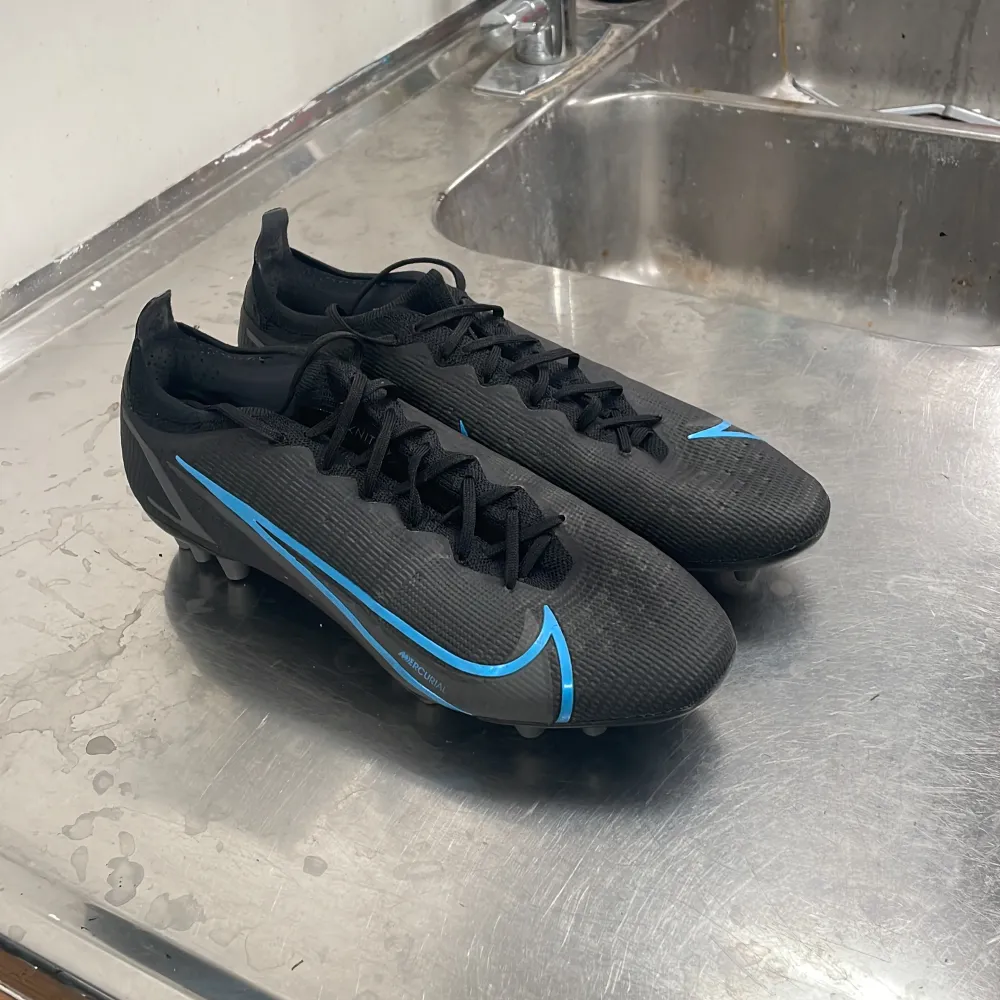 Säljer ett par svarta Nike fotbollsskor i syntetmaterial med snygga blå detaljer. Skorna har snörning och är i mycket bra skick. Perfekta för fotbollsträning eller match. De har en tight passform och är designade för att ge bra grepp på planen.. Skor.