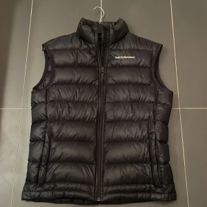 Marinblå/svart dunväst från Peak Performance - Säljer en svart dunväst från Peak Performance i storlek M. Västen har en dragkedja framtill och är perfekt för kyliga dagar. Den är i mycket bra skick och passar både höst och vinter. Perfekt för lager-på-lager-stil! 