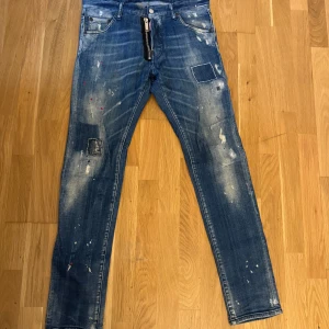 Blå jeans från DSQUARED2 - Säljer ett par snygga blå jeans från DSQUARED2 i storlek 46. De har en cool distressed look med slitningar och färgstänk. Perfekta för en avslappnad och trendig stil. Midjan är midwaist och passformen är slim. Perfekta för både vardag och fest!