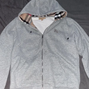 Grå burberry - Säljer en snygg grå huvtröja från Burberry i mycket bra skick. Den har en dragkedja framtill och en klassisk rutig detalj i huvan. Perfekt för höst och vår, med en normal passform och långa ärmar. Superbekväm och stilren!