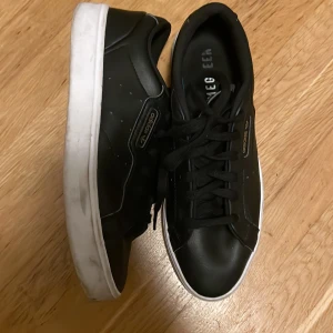 Svarta Adidas skor - Säljer ett par svarta Adidas skor i skinn med klassisk design. De har en vit sula och snörning framtill. Perfekta för vardagsbruk och ger en stilren look. Lite slitna på sulan men annars i bra skick.