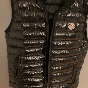 Svart dunväst från Moncler - Säljer en snygg svart dunväst från Moncler i mycket bra skick. Västen har en glansig yta. Perfekt för höst och vår, med klassisk Moncler-logga på bröstet. Den är både stilren och praktisk för kyligare dagar. Sparsamt använd. Storlek medium.