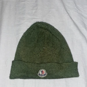Grön ribbad mössa från Moncler - Säljer en snygg grön ribbad mössa från Moncler. Perfekt för kyliga dagar med sin stilrena design och varma material. Den har en uppvikt kant och Moncler-loggan framtill. Passar både till vardags och utomhusaktiviteter.