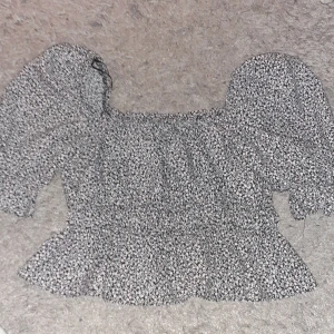 Blommig topp från Divided - Säljer en supersöt blommig topp från Divided i storlek S. Den har en svart bas med små rosa och vita blommor. Toppen har puffiga trekvartsärmar och en volangdetalj längst ner. Perfekt för vår och sommar och födelsedagar!