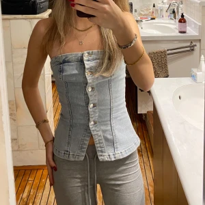 Jeans tubtopp med knappar - Snygg jeans tubtopp med knappar framtill. Perfekt för en avslappnad sommarlook eller en utekväll. Toppen är ärmlös och har en tight passform som framhäver figuren. Den ljusblå denimfärgen ger en klassisk touch.