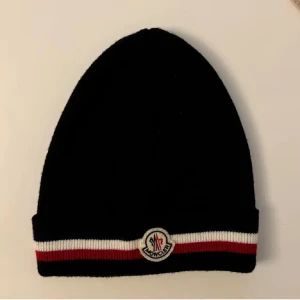 Svart mössa med Moncler-logga - Snygg svart mössa från Moncler med en klassisk design. Den har en ribbad kant i rött och vitt samt en broderad Moncler-logga framtill. Perfekt för att hålla sig varm under kyliga dagar.