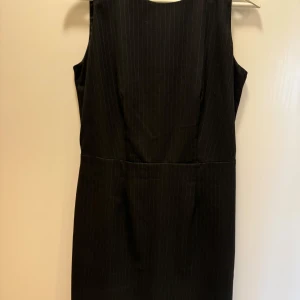 Svart ärmlös klänning från Sinéquanone - Elegant svart ärmlös klänning från Sinéquanone i ett klassiskt snitt. Perfekt för både kontoret och festliga tillfällen. Klänningen har en diskret randig textur som ger en sofistikerad touch. Den är i mycket bra skick och passar utmärkt för höst och vår.