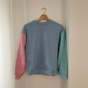 Pastellfärgad sweatshirt - Säljer en superfin pastellfärgad sweatshirt med en cool färgblock-design. Tröjan har en ljusblå bas med en rosa ärm och en mintgrön ärm. Perfekt för en avslappnad stil och passar bra till både jeans och kjol. Den är i bra skick och redo för nya äventyr!