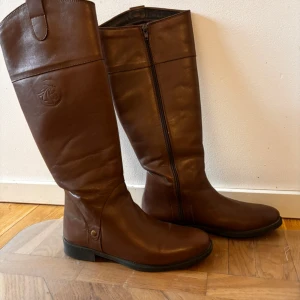 Bruna ridstövlar i skinn - Snygga bruna ridstövlar i skinn med dragkedja på sidan. Perfekta för höst och vinter, dessa stövlar har en klassisk design med en liten klack och rundad tå. De är i mycket bra skick.  Passar perfekt för ridning eller en stilren vardagslook.