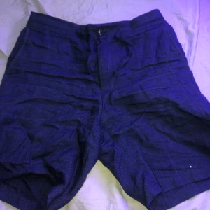 Mörkblå shorts från These Glory Days - Säljer ett par snygga mörkblå shorts som aldrig är använda från These Glory Days i storlek 44. De är tillverkade i linne vilket gör dem perfekta för varma sommardagar. Shortsen har en elastisk midja och knappar bak för en stilren look. Perfekta för både vardag och fest! 