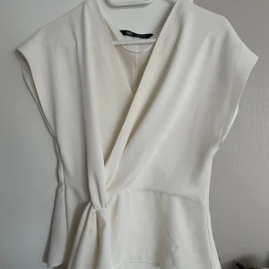 Vit omlottopp från Zara - Säljer en trendig vit blus från Zara i storlek M. Blusen har en elegant knytdetalj i midjan och är ärmlös, vilket gör den perfekt för både vardag och fest. Materialet känns mjukt och bekvämt mot huden. Passar utmärkt till både jeans och kjol! Lite nopprig på vänstra nedre delen, men märks inte av så mycket. Nypris var 350kr men säljer för ynka 150kr!