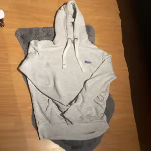 Säljer en snygg grå hoodie från Superdry i bra skick. Den har en klassisk design med justerbar huva och en liten broderad logga på bröstet. Perfekt för både höst och vår, Superbekväm och stilren!