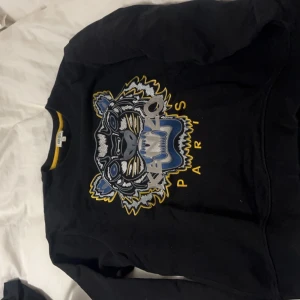 Svart sweatshirt med tigertryck från Kenzo - Säljer en svart sweatshirt från Kenzo med ett coolt tigertryck i blått, gult och grått. Tröjan har långa ärmar och en rund halsringning med en gul randdetalj. Perfekt för en avslappnad och stilren look. Passar både till vardags och fest!