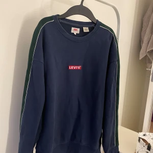 Mörkblå sweatshirt från Levi's - Säljer en snygg mörkblå sweatshirt från Levi's i storlek M. Tröjan har vita och gröna ränder längs ärmarna och det klassiska Levi's-loggan på bröstet. Perfekt för en avslappnad stil och passar bra till både jeans och sneakers.