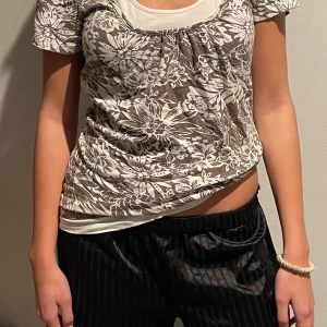 Blommig kortärmad topp - Säljer en söt kortärmad topp med blommigt mönster i brunt och vitt. Toppen har en rundad halsringning med en vit kant som ger en fin kontrast. Perfekt för en avslappnad sommarlook!