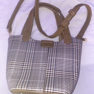 Rutig axelväska från Chrisbella - Säljer en snygg rutig axelväska från Chrisbella i mycket bra skick. Väskan har ett klassiskt rutigt mönster i svart och beige med bruna detaljer och axelrem. Perfekt för både vardag och fest!