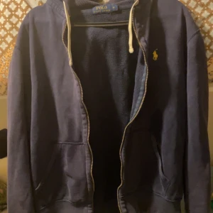 Mörkblå hoodie från Polo Ralph Lauren - Säljer en snygg mörkblå hoodie från Polo Ralph Lauren i storlek M. Den har en dragkedja framtill och en liten broderad logga på bröstet. Perfekt för höst och vår, med en skön passform och långa ärmar. Huvan har justerbara snören för extra komfort. Passar både till vardags och lite mer avslappnade tillfällen.