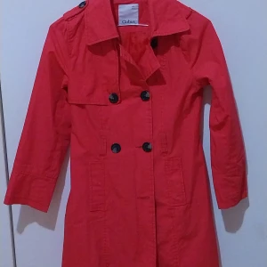 Röd trenchcoat från Cubus - Säljer en snygg röd trenchcoat från Cubus i storlek 140. Den har långa ärmar och knappar framtill. Perfekt för höst och vår, med en klassisk stil som funkar till många tillfällen. Jackan är i bra skick och redo för nya äventyr!
