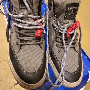 Grå och svarta sneakers från S.Oliver - Snygga och robusta sneakers från S.Oliver i grått och svart. De har en cool design med snörning och en bekväm sula. Perfekta för höst och vår, med en stilren look som passar både till vardags och lite mer uppklädda tillfällen.