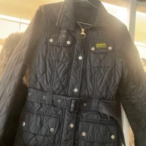 Svart quiltad jacka från Barbour - Säljer en snygg svart quiltad jacka från Barbour i storlek 34. Jackan har ett bälte i midjan och flera fickor med knappar. Perfekt för höst och vår med sin stilrena design och praktiska detaljer. Passar både till vardags och lite finare tillfällen. 🖤
