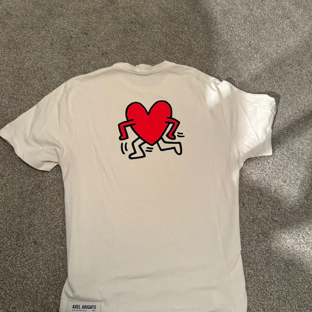 Säljer en vit t-shirt från Axel Arigato med tryck fram och bak. På framsidan står det 'ARIGATO ©2014' och 'Keith Haring' i rött. Baksidan har ett coolt rött hjärta med ben. Perfekt för en casual look!. T-shirts.