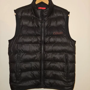 Hugo Boss väst - Säljer en stilren Hugo Boss-väst i nyskick. Elegant svart färg, quiltad design och praktiska fickor med dragkedja. Perfekt för lager-på-lager eller som ytterplagg på svala dagar. Passar både casual och sportiga outfits!