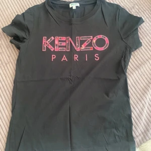 Svart t-shirt från Kenzo - Säljer en snygg svart t-shirt från Kenzo med rosa logga på framsidan. Den är i storlek S och gjord av 100% bomull, vilket gör den superbekväm. Perfekt för vardagsbruk eller en avslappnad utekväll. T-shirten har korta ärmar och en klassisk passform.