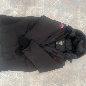 Svart dunjacka från Canada Goose - Säljer en svart dunjacka från Canada Goose i storlek XL. Jackan är perfekt för kalla vinterdagar med sin tjocka vaddering och varma huva. Den har en dragkedja framtill och en snygg röd lönnlövsdetalj. Märkets ikoniska logga syns på ärmen. Passar perfekt för vinterns alla äventyr! 