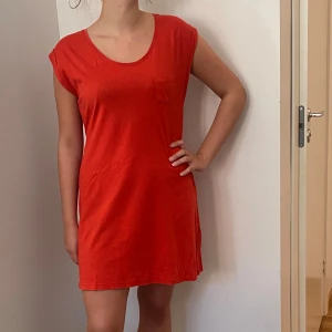 Röd kortklänning med knytband - Säljer en snygg röd kortklänning med ett praktiskt knytband i midjan. Klänningen har en enkel design med korta ärmar och en liten bröstficka. Perfekt för sommardagar eller en avslappnad fest. Materialet känns mjukt och bekvämt mot huden. Uppskattad strl M