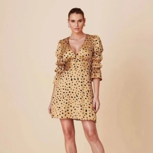 Adoore mini leopard klänning XS - En vacker klänning med prickigt mönster i beige och svart från Adoore. Klänningen har en V-ringning och är figursydd. Den har också en knappstängning i fram och är perfekt för en festlig tillställning.   OBS oanvänd med etiketter!