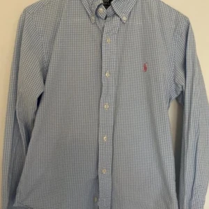 Blå rutig skjorta från Polo Ralph Lauren - Säljer en snygg blå rutig skjorta från Polo Ralph Lauren i storlek S. Skjortan är slim fit och har långa ärmar. Den är tillverkad i bomull och har en klassisk krage med knappar. Det finns en broderad logga i rött på bröstet. Perfekt för både vardag och fest!
