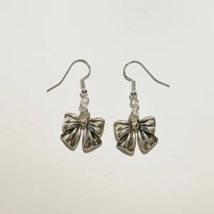 Söta silverörhängen med rosett - Säljer ett par supersöta handgjorda örhängen i silverfärg med en fin rosettdetalj. Perfekta för att ge en gullig touch till vilken outfit som helst. Passar perfekt till både vardag och fest!